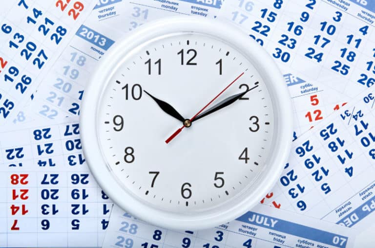 An analog clock lies on top of a stack of paper calendars.